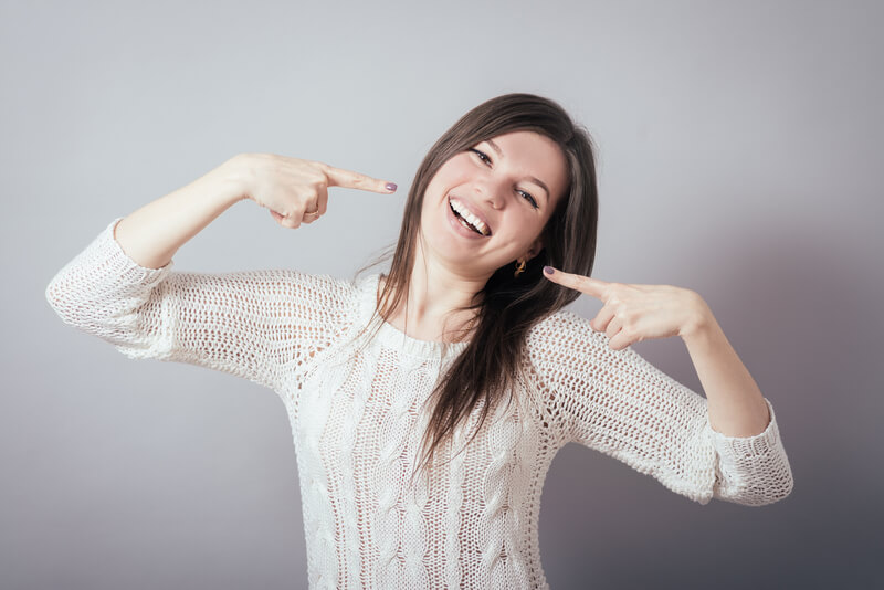 woman in white sweater pointing at her face with both hands and laughing